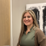 A woman with long, straight hair wearing a green shirt and a brown cardigan smiles at the camera. She is positioned beside a modern artistic background featuring black and white designs.