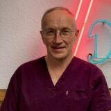 A middle-aged man wearing purple scrubs stands in front of a neon sign. He has short hair and glasses, smiling softly at the camera.