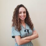 A smiling woman with curly hair stands confidently with her arms crossed, wearing a light blue outfit. The background is a plain, neutral color.