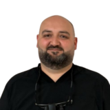 A person with a beard is wearing a black shirt and dental loupes around their neck, standing against a white background.
