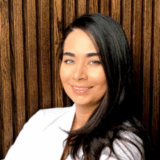 A smiling young woman with long, dark hair poses against a textured wooden background. She wears a white top and has striking green eyes, conveying a friendly and approachable demeanor.