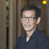 A man with short black hair and glasses smiles while sitting against a softly lit background of blurred vertical lines. He is wearing a dark shirt and appears approachable and confident.