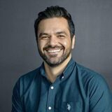 A smiling man with dark hair and a beard wears a blue shirt and stands against a grey background. His expression conveys warmth and approachability.