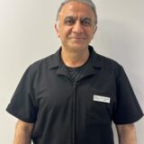 A professional man wearing a black uniform shirt stands against a plain background, smiling. He appears confident and approachable, with neatly combed hair and a name tag on his left side.