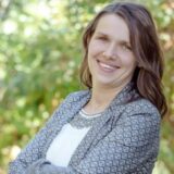 A woman with long brown hair smiles confidently, standing with her arms crossed in front of a blurred green background. She wears a patterned blazer over a white shirt and exudes a friendly demeanor.