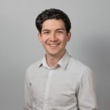 A young man with dark, curly hair smiles warmly. He is wearing a light-colored button-up shirt and stands against a neutral gray background.