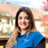 A woman with long, dark hair smiles while standing outdoors, wearing a blue shirt with an embroidered name. Soft-focus trees with autumn foliage are visible in the background, creating a warm, inviting atmosphere.