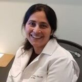 A smiling woman in a white lab coat sits at a desk, looking directly at the camera. Her dark hair is styled down, and the background features a neutral wall.