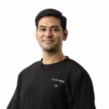 A smiling male doctor wearing a black medical scrub top, looking confidently at the camera against a plain white background.