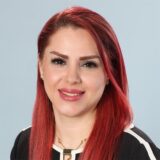 A woman with long, bright red hair smiles at the camera, wearing a black and white striped top. The background is a light blue, providing a clean and professional look.