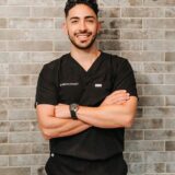 A smiling male medical professional wearing black scrubs stands confidently with his arms crossed against a textured gray wall.