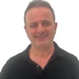 A middle-aged man with short dark hair and a slight smile is wearing a black shirt against a plain white background.