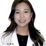 A smiling woman wearing a white lab coat, identified as Dr. M. Su, with long dark hair. She appears confident and approachable, set against a plain white background.