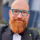 A smiling man with a bald head and a vibrant red beard wears a black tuxedo with a bow tie. He has blue-framed glasses and is posing outdoors with a blurred background.