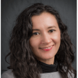 A woman with long, curly hair smiles at the camera. She wears a grey sweater and a black turtleneck, set against a dark background.