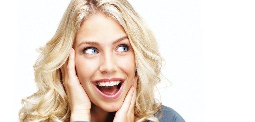 Une jeune femme aux longs cheveux blonds ondulés affiche une expression enthousiaste, les mains sur les joues et un sourire éclatant.