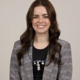 A smiling woman with long brown hair stands against a neutral background. She wears a checked blazer over a black top, accented by a necklace.