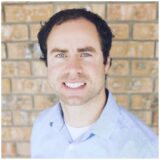 A smiling man with short dark hair and blue eyes, wearing a light blue button-up shirt, stands in front of a textured brick wall.