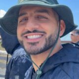 A smiling man with a beard and mustache wears a green bucket hat. He is outdoors, with a clear blue sky in the background, and is dressed in a dark jacket. His bright expression conveys a cheerful atmosphere.