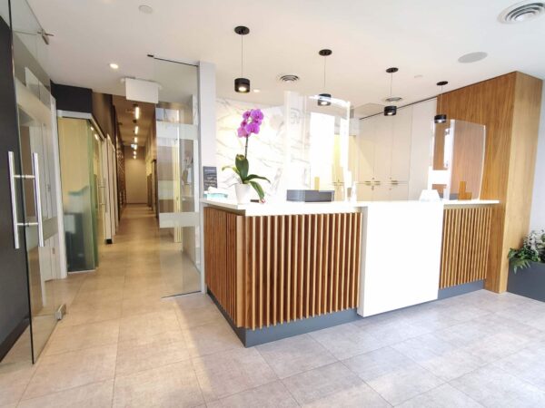 A modern reception area featuring a wooden front desk with an orchid, surrounded by sleek glass partitions and stylish lighting. The space is bright and inviting, with tiled flooring and minimalist decor.