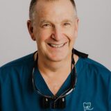 A smiling male veterinarian wearing a teal medical scrubs top and dental loupes around his neck, posing against a plain background.