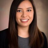A smiling woman with long, dark hair, wearing a black blazer over a grey top, is portrayed against a soft, neutral background.