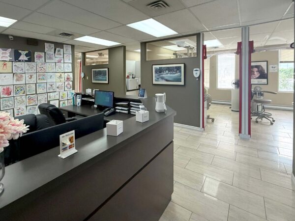 A modern office reception area featuring a dark reception desk with flowers, a wall adorned with colorful artwork, and a view into a treatment area with dental chairs and equipment. Brightly lit with natural light, the space conveys a welcoming atmosphere.