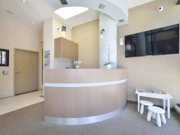 A modern reception area featuring a curved wooden desk, a flat-screen TV on the wall, and minimalistic seating. The space is bright and inviting, with light-colored walls and a clean, organized appearance.
