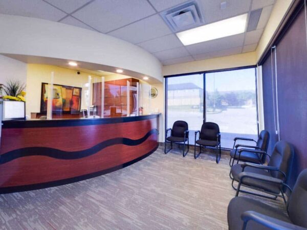 A modern reception area featuring a curved wooden front desk, multiple chairs arranged for waiting visitors, and large windows allowing natural light. The walls are painted in soft colors, creating a welcoming atmosphere.