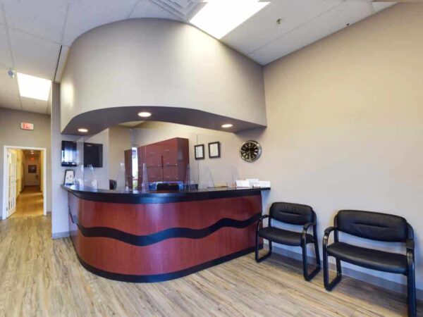 A modern reception area featuring a curved front desk, wooden paneling, and two black chairs. The walls are painted a neutral color, and there are framed pictures and a clock on the wall, creating a welcoming atmosphere.