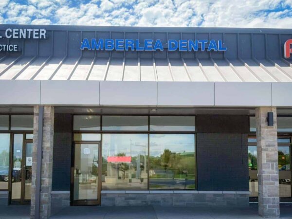 A dental clinic named "Amberlea Dental" is prominently displayed on a modern storefront, featuring large windows and a sleek metal roof. The surrounding area includes other businesses, with a clear blue sky overhead.
