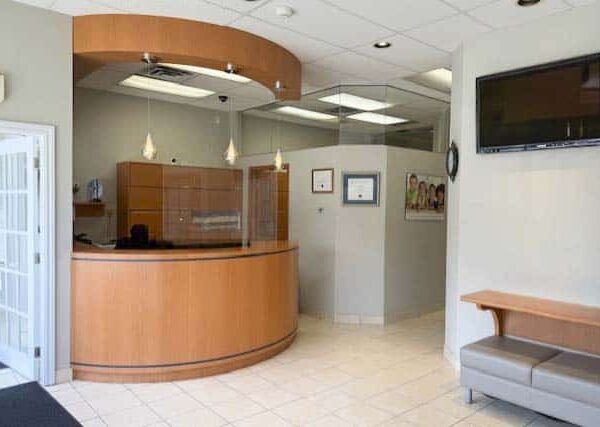 A clean, modern reception area featuring a wooden front desk, light-colored walls, and tiled flooring. There’s a waiting bench and a wall-mounted television, with natural light coming through windows.