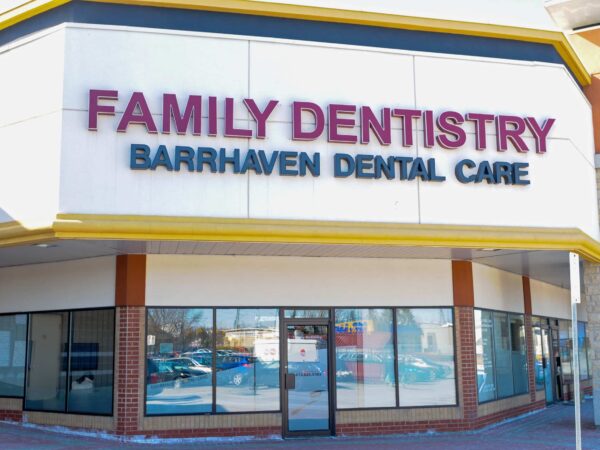 The image shows the exterior of a dental clinic named "Family Dentistry" with the additional text "Barrhaven Dental Care." The building features large windows and a bright, welcoming sign.