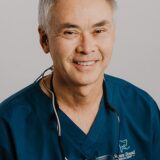 A smiling male dentist wearing blue scrubs and dental loupes. The background is neutral, highlighting his professional demeanor.