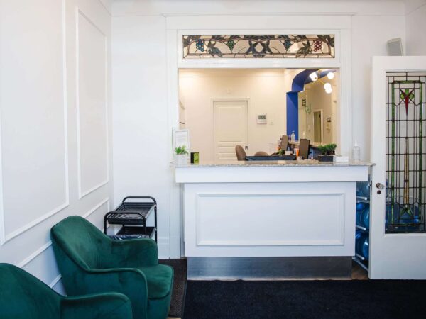 A welcoming reception area featuring a white counter, two green chairs, and a door with glass panels. The background shows a bright, inviting space with soft lighting and minimal decor, suggesting a calm environment.