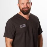A smiling man in a dark medical scrub top stands confidently against a plain background. He has a beard and short hair, wearing an orange wristwatch, and is likely a healthcare professional.