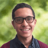 A smiling young man with glasses, wearing a blue shirt and a maroon cardigan, is photographed against a blurred green background, suggesting a natural outdoor setting.