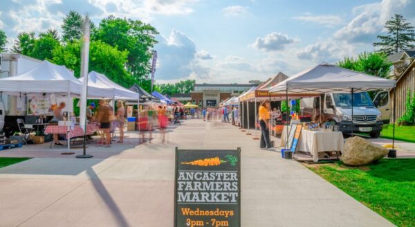 A sunny outdoor farmers market scene with several white tents set up along a walkway. A sign indicates "Ancaster Farmers Market" and its hours, with vendors showcasing various goods. Green trees and fluffy clouds form the backdrop.