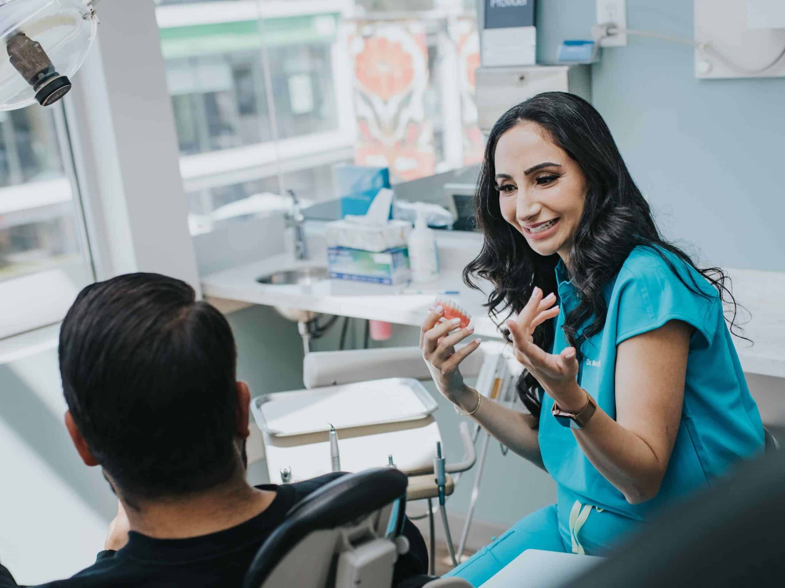 A dental professional in a blue outfit is engaging in a friendly conversation with a patient seated in a dental chair. The bright and inviting clinic features natural light, creating a warm atmosphere, while tools and equipment are visible in the background. The 123Dentist Smiley adds a cheerful touch.