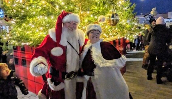 A cheerful Santa Claus and a woman dressed in a festive outfit pose together in front of a beautifully decorated Christmas tree, surrounded by holiday lights and a crowd of people enjoying the festive atmosphere.