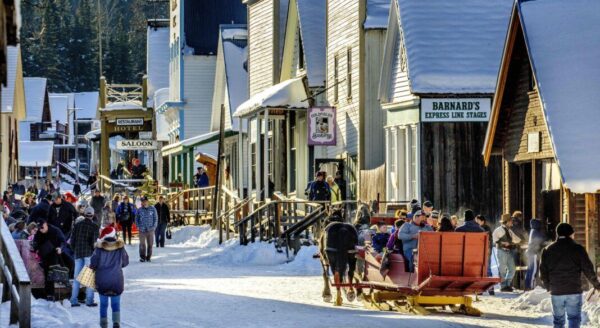 A bustling winter street scene with snow-covered buildings and people shopping, walking, and enjoying outdoor activities. Sleds are present, and the atmosphere captures a lively, festive spirit in a historic town.