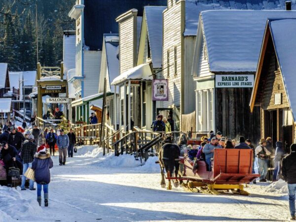 A bustling winter street scene with snow-covered buildings and people shopping, walking, and enjoying outdoor activities. Sleds are present, and the atmosphere captures a lively, festive spirit in a historic town.