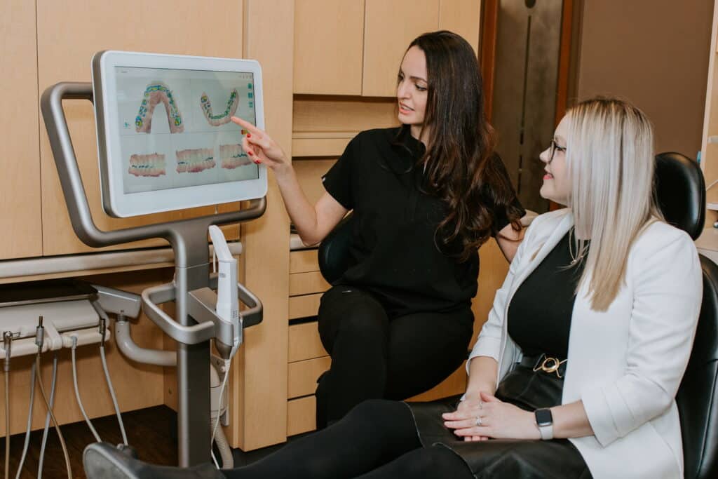 A dental professional discusses treatment options with a patient, pointing at visual information on a digital screen in a modern dental office. The patient sits comfortably, engaged in the conversation.