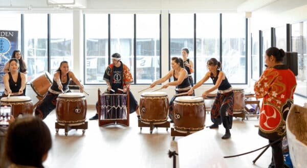 A group of performers plays traditional drums in a spacious room filled with natural light. The musicians wear vibrant outfits and focus on their rhythmic performance, while an audience observes from the background.
