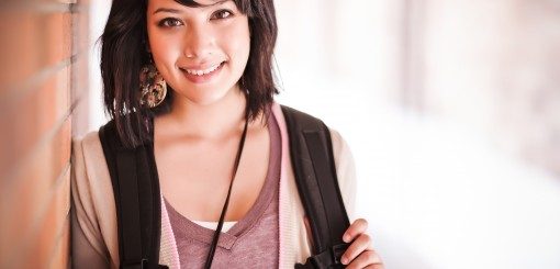 A young woman with short dark hair smiles while leaning against a wall, holding a backpack strap. She is dressed casually and appears friendly in a softly lit environment.