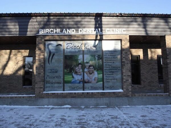 Exterior of a dental clinic with a brick facade. Large front window displays an image of smiling people and dental graphics. The clinic's name is visible above the entrance. Snow is on the ground, indicating a cold season.