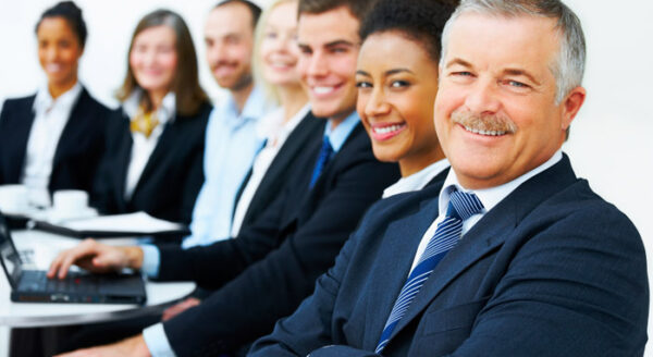 A diverse group of professionals in business attire smiles while seated in a row, with a laptop visible in front of one person. The setting suggests a collaborative work environment or a meeting.