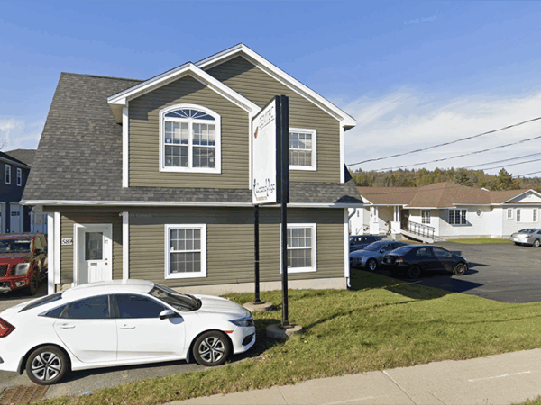 A two-story building with a gray exterior and peaked roof sits beside a parking lot filled with various vehicles. A white car is parked in front, with power lines and clear blue skies in the background.