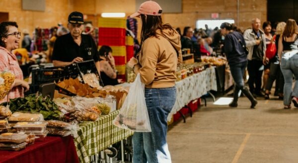 A busy indoor market scene featuring shoppers browsing various stands. A person in casual clothing holds a shopping bag while inspecting items on a table. Vendors interact with customers in a vibrant, bustling atmosphere filled with colorful displays of food and goods.