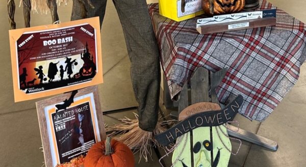 A festive Halloween display featuring a scarecrow wearing plaid pants, next to a table adorned with pumpkins, decorations, and flyers for a Halloween event. The scene captures the spirit of autumn and Halloween festivities.
